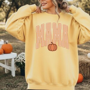 Pode incluir: Uma camisola de manga comprida e gola redonda em amarelo manteiga com a palavra MAMA impressa num padr&atilde;o de xadrez vichy laranja e branco. Um pequeno gr&aacute;fico de uma ab&oacute;bora laranja com bolinhas brancas est&aacute; centrado sob a palavra MAMA.