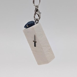 May include: A small, rectangular pendant with a black cord. The pendant is off-white with a small black dagger design. A silver clasp and a dark blue accent are also visible.