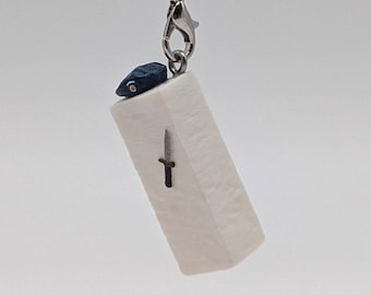 May include: A small, rectangular pendant with a black cord. The pendant is off-white with a small black dagger design. A silver clasp and a dark blue accent are also visible.