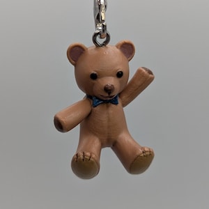 May include: A small brown teddy bear charm with a dark blue bowtie, attached to a silver-toned metal lobster clasp and a black cord loop. The bear has black eyes, a stitched nose, and textured paw pads on its feet, suspended by a metal ring.