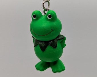 May include: A green frog keychain with a black collar and white eyes. The frog is smiling and attached to a silver metal chain and keyring. The background is a solid light gray.
