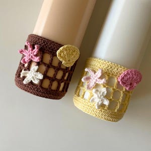 May include: Two crochet bottle cozies, one brown and one yellow, each decorated with floral and seashell details. The brown cozy has pink and white flowers, and the yellow one has pink and white flowers and a yellow seashell.