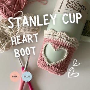 Puede incluir: Un cubre vaso de crochet de color rosa y blanco en forma de corazón con una abertura en forma de corazón. El cubre vaso está sobre un vaso Stanley Quencher H2O Tumbler azul claro. El texto "STANLEY CUP" es visible en el vaso. Dos agujas de crochet son visibles en la parte inferior de la imagen, una rosa y otra azul. El texto "PINK" y "BLUE" está escrito en las agujas.