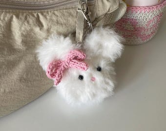 May include: A fluffy white bunny keychain with a pink crocheted bow. The bunny has black eyes and a pink nose. The keychain is attached to a beige bag with a silver zipper.