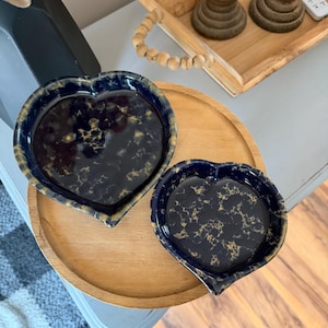 May include: Two heart-shaped dishes with a glossy dark navy blue finish featuring a mottled gold and tan marbled pattern. The dishes have raised edges and a smooth, reflective surface, showcasing a distinct heart silhouette in two different sizes.