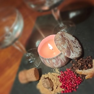 May include: A small, round candle in a tin container with a floral design. The candle is lit and the flame is visible. The candle is surrounded by various spices, including cloves, peppercorns, and cinnamon sticks.