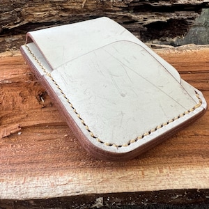Puede incluir: Una cartera de cuero blanco con detalles de costura marrón. La cartera tiene un cierre de solapa y un solo bolsillo.