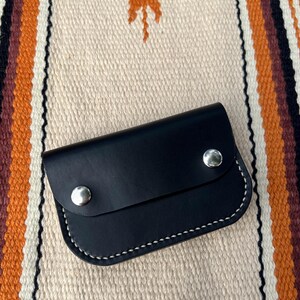 May include: A black leather wallet with a flap closure and two silver snaps. The wallet is on a white and orange striped background.