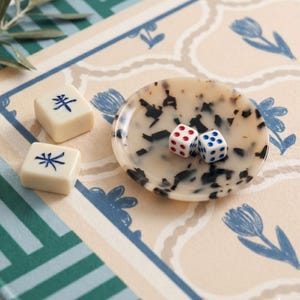 May include: A close-up shot of game pieces and dice on a patterned surface. Two white dice with red and blue dots sit in a small, speckled dish. Two ivory-colored tiles with blue symbols are also present, suggesting a game like Mahjong.