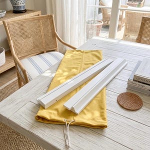 May include: Two white wooden display ledges with a grooved front edge resting on a folded mustard yellow fabric drawstring bag. The items are positioned on a white wooden table next to a stack of two books and a small round woven coaster.