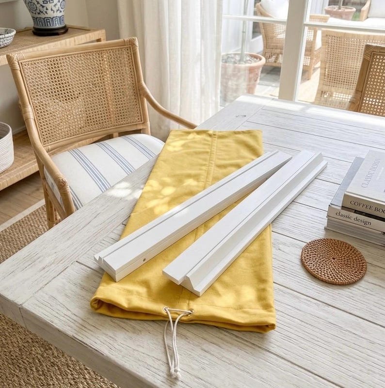 May include: Two white wooden display ledges with a grooved front edge resting on a folded mustard yellow fabric drawstring bag. The items are positioned on a white wooden table next to a stack of two books and a small round woven coaster.