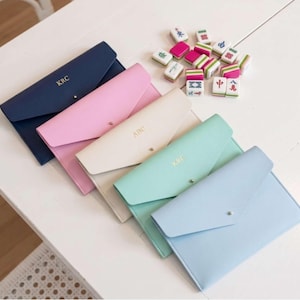 Op de afbeelding: Een assortiment gepersonaliseerde leren enveloppe-achtige etuis in marineblauw, roze, crème, mintgroen en lichtblauw. Elke etui heeft een druksluiting en gouden letters. Een verzameling kleurrijke Mahjong-tegels ligt in de buurt.