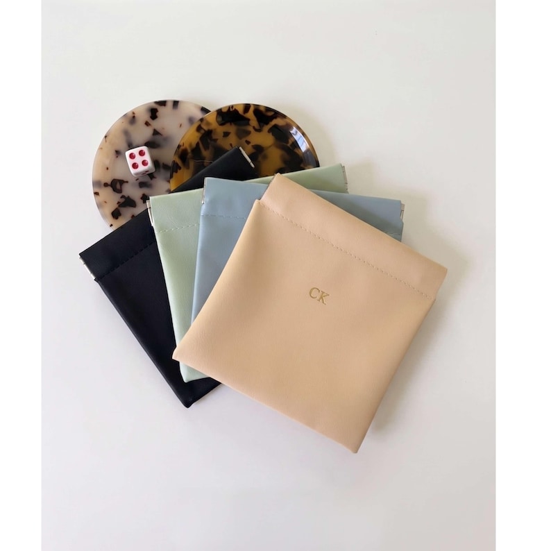 May include: An assortment of small, square pouches in black, light green, and light blue, with a tan pouch featuring the initials "CK". Two decorative coasters with a tortoiseshell pattern and a dice are also present.