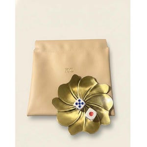 Puede incluir: Un cuenco para baratijas en forma de flor dorada con dos dados y una bolsa beige. Los dados son blancos con puntos azules y rojos. La bolsa tiene las iniciales "KC" en relieve. El fondo es de color beige claro.