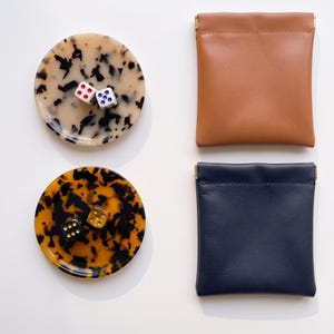 Puede incluir: Dos bandejas circulares con patrones de carey, una crema y otra ámbar, cada una con dos dados. Junto a ellas hay dos bolsas cuadradas, una color canela y otra azul marino, con cierres de marco metálico.