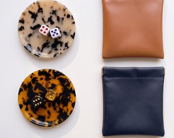 May include: Two circular trays with tortoiseshell patterns, one cream and one amber, each holding two dice. Beside them are two square pouches, one tan and one navy blue, featuring metal frame closures at the top edges.