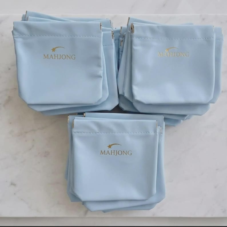 May include: A collection of light blue pouches with the word "MAHJONG" printed in gold. Each pouch has a drawstring closure and a small star design above the word. The pouches are arranged in a pyramid shape.