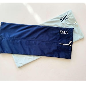 Puede incluir: Dos fundas rectangulares de tela, una azul marino y otra azul claro. La funda azul marino tiene bordado blanco "KMA". La funda azul claro tiene bordado negro "KRC". Ambas tienen cierre de cordón.