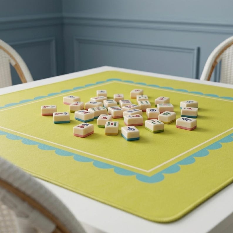 K&ouml;nnte beinhalten: Ein Satz elfenbeinfarbener Mahjong-Steine mit farbenfrohen Markierungen liegt auf einer limettengr&uuml;nen Matte. Die Matte hat einen wellenf&ouml;rmigen Rand in Hellblau. Die Steine haben rote, blaue und gr&uuml;ne Akzente. Das Spiel steht auf einem wei&szlig;en Tisch.