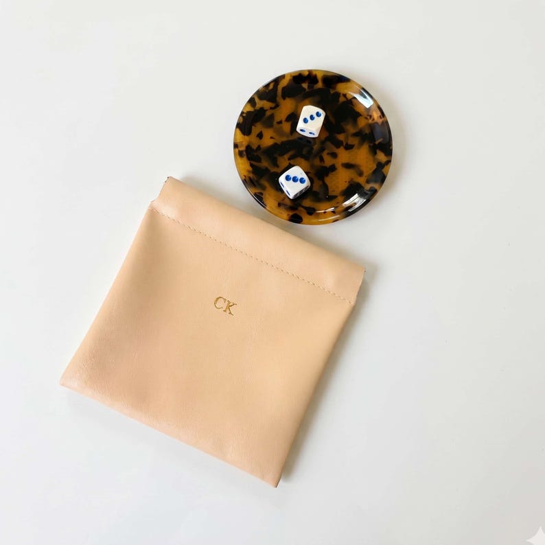 May include: A tan leather pouch with the initials "CK" in gold lettering, next to a tortoiseshell-patterned dish holding two white dice with blue dots. The dice show a 2 and a 5.