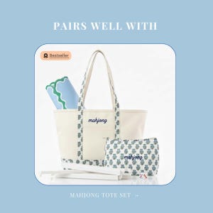 May include: A cream-colored canvas tote bag and a matching zippered pouch, both embroidered with the word mahjong in blue script. The set features a floral pattern on the straps and base, accompanied by a blue and green mat and white game sticks.