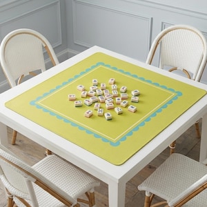 K&ouml;nnte beinhalten: Ein quadratischer wei&szlig;er Tisch mit vier wei&szlig;en Korbst&uuml;hlen. Eine gr&uuml;ne Matte mit einem blau geschwungenen Rand und einem wei&szlig;en Innenrand bedeckt den Tisch. Mahjong-Steine sind auf der Matte verstreut.
