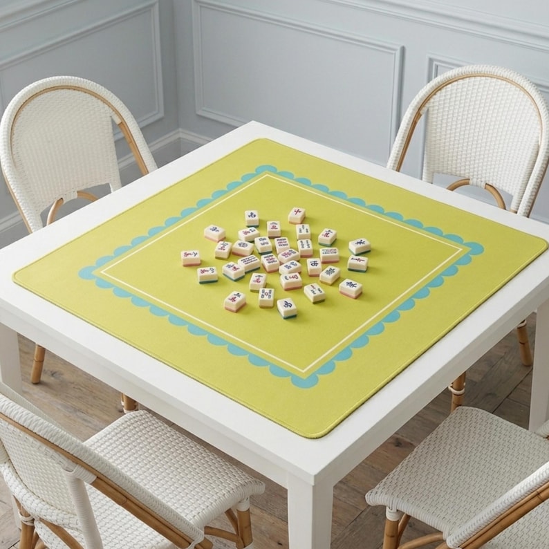 K&ouml;nnte beinhalten: Ein quadratischer wei&szlig;er Tisch mit vier wei&szlig;en Korbst&uuml;hlen. Eine gr&uuml;ne Matte mit einem blau geschwungenen Rand und einem wei&szlig;en Innenrand bedeckt den Tisch. Mahjong-Steine sind auf der Matte verstreut.