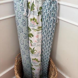 May include: Three tall, cylindrical fabric storage bags with drawstring closures, standing upright in a woven basket. The bags feature distinct patterns: one with a light blue floral print, one with a green and pink botanical design, and one with a dark teal leaf motif.