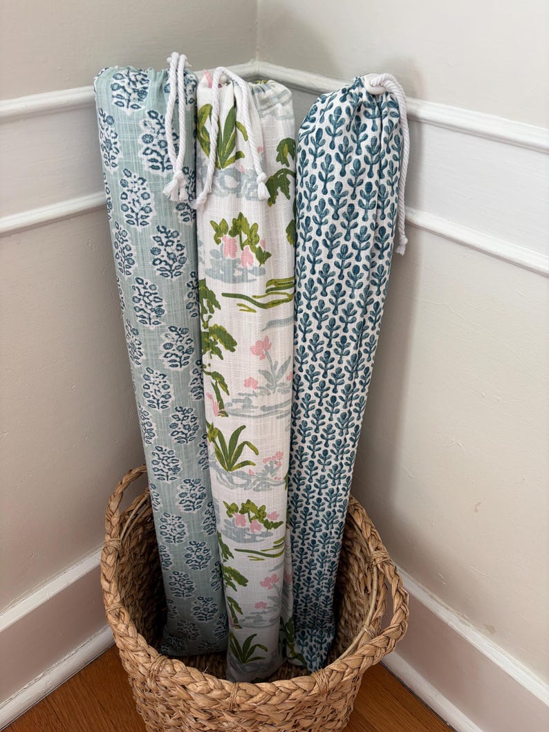 May include: Three tall, cylindrical fabric storage bags with drawstring closures, standing upright in a woven basket. The bags feature distinct patterns: one with a light blue floral print, one with a green and pink botanical design, and one with a dark teal leaf motif.