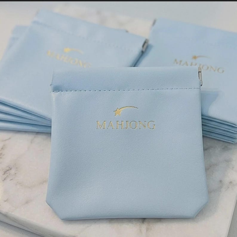May include: Several light blue pouches with the word "MAHJONG" printed in gold. Each pouch has a star and curved line design above the word. The pouches are made of a soft material and are likely used for storing small items.