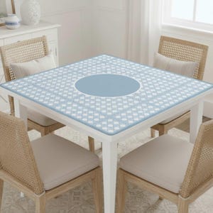 Pu&ograve; includere: Un tavolo quadrato bianco con un piano a motivi blu chiaro e bianco. Il tavolo ha un cerchio blu centrale ed &egrave; circondato da quattro sedie in vimini con cuscini beige. La stanza &egrave; ben illuminata con una finestra sullo sfondo.