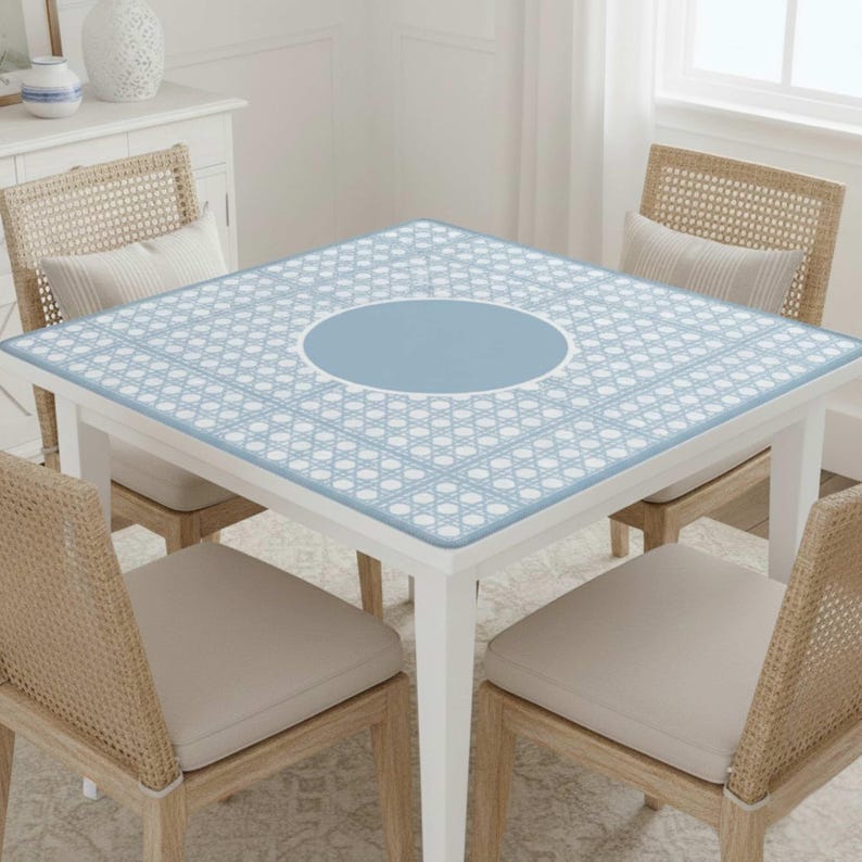 Pu&ograve; includere: Un tavolo quadrato bianco con un piano a motivi blu chiaro e bianco. Il tavolo ha un cerchio blu centrale ed &egrave; circondato da quattro sedie in vimini con cuscini beige. La stanza &egrave; ben illuminata con una finestra sullo sfondo.