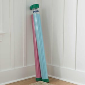 May include: Two long, cylindrical pool noodles, one pink and one light blue, lean against a white wall. The top of the noodles have a green cap. The text "MHB" is printed on the top of the blue noodle. The noodles rest on a beige rug.
