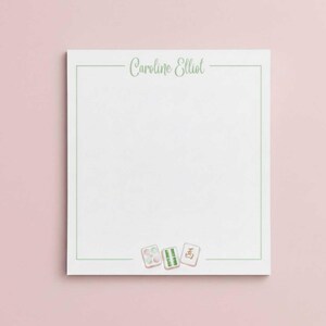 May include: White notepad with a green border and the name "Caroline Elliot" in a script font. Three Mahjong tiles are at the bottom. The background is a soft pink.