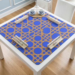 May include: A square blue Mahjong table mat with a gold geometric pattern. Four white tile racks are placed around the mat, with Mahjong tiles inside. Two teacups and a water carafe are on the table.