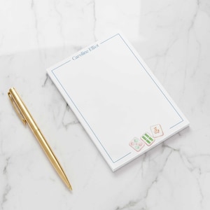 May include: A white notepad with a blue border and the name "Caroline Elliot" printed at the top. The notepad has a decorative design of Mahjong tiles at the bottom and a gold pen.