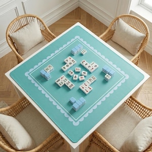 Puede incluir: Una mesa cuadrada blanca con un tapete de juego turquesa con un borde festoneado. El tapete muestra un juego de Mahjong en progreso, con fichas blancas y soportes de fichas azules. Cuatro sillas de mimbre con cojines beige rodean la mesa.