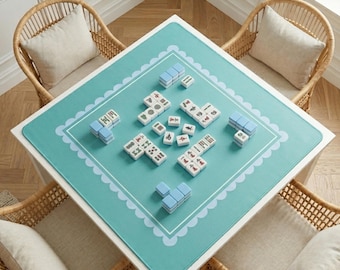 Puede incluir: Un tapete de mahjong cuadrado de color verde azulado claro con un borde festoneado blanco, colocado sobre una mesa blanca. El tapete presenta varios grupos de fichas de mahjong blancas y azul claro dispuestas en diversas formaciones.