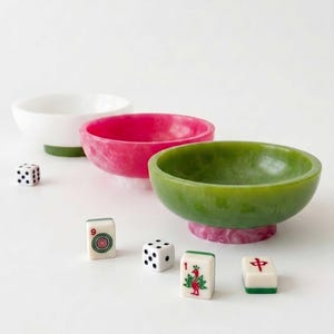 May include: Three small, round bowls with a marbled finish in white, pink, and green, each featuring a contrasting coloured base. Scattered around the bowls are two six-sided white dice and three rectangular mahjong tiles with red and green designs.