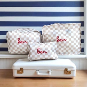 May include: A collection of three beige and white chequered bags and a white suitcase. The bags are embroidered with the name "ben" in red. The suitcase has gold-coloured hardware and a white handle. The background features blue and white striped wallpaper.