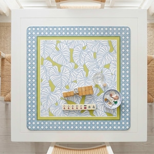 May include: Overhead view of a white square table with a patterned mat featuring a leaf design in white and blue on a green background. A small bowl with shells and dice, a glass, and mahjong tiles are on the mat.