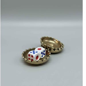 May include: Two small, gold-colored scalloped dishes. One dish contains two white dice with red and blue dots. The other dish is empty. The dishes are set against a light gray background.