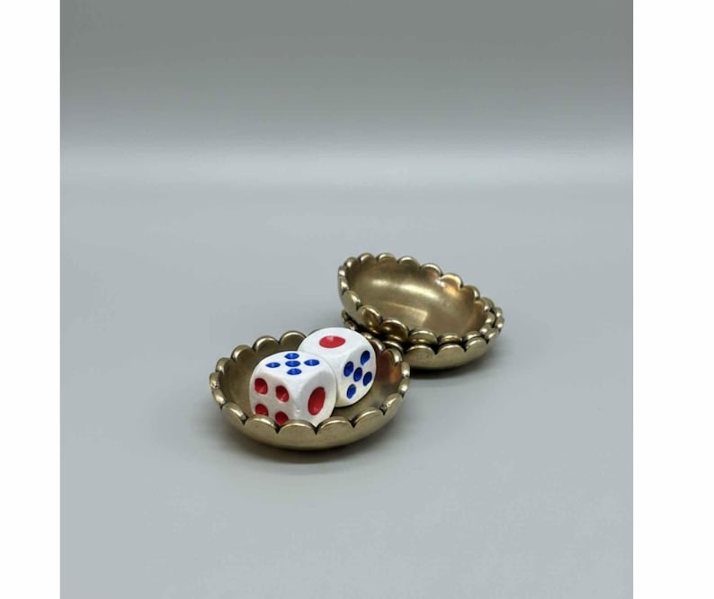 May include: Two small, gold-colored scalloped dishes. One dish contains two white dice with red and blue dots. The other dish is empty. The dishes are set against a light gray background.