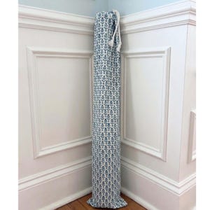 May include: A tall, cylindrical yoga mat bag made of fabric with a blue and white leaf pattern. The bag is closed at the top with a drawstring and tassel. The bag is standing upright in the corner of a room with white wainscoting.