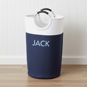 May include: A cylindrical laundry hamper with a navy blue lower section and a white upper section. The word "JACK" is printed in light blue on the navy section. It has two black and silver handles.