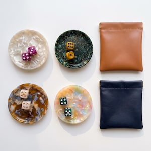 May include: Four circular resin trays with marbled patterns in white, dark green, brown, and iridescent tones, each containing two six-sided dice. Two rectangular pouches in tan and navy blue leather are positioned to the right of the trays.