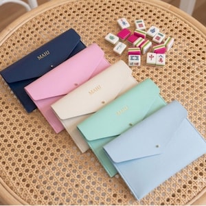 May include: Five rectangular envelope-style pouches in navy, pink, cream, mint green, and light blue, each featuring a gold-toned snap closure and the gold-embossed text MAHJ. A small collection of colorful mahjong tiles is arranged nearby.