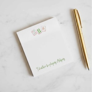 May include: White notepad with watercolor Mahjong tiles and the text "I'd rather be playing Mahjong" in green. A gold pen rests on the right side of the notepad, on a white marble surface.