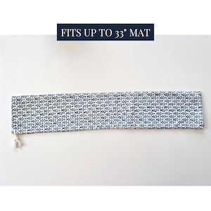 May include: A long, rectangular fabric yoga mat bag featuring a repeating blue and white floral pattern. The bag includes a white drawstring closure at one end and text above the bag that reads FITS UP TO 33" MAT in white on a dark blue background.