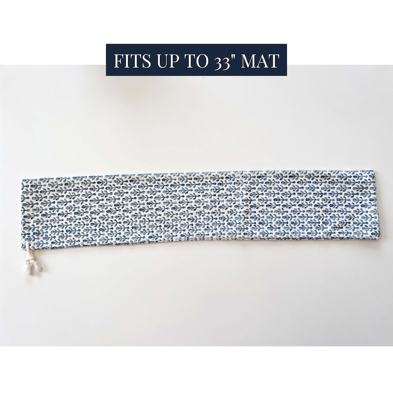 May include: A long, rectangular fabric yoga mat bag featuring a repeating blue and white floral pattern. The bag includes a white drawstring closure at one end and text above the bag that reads FITS UP TO 33" MAT in white on a dark blue background.